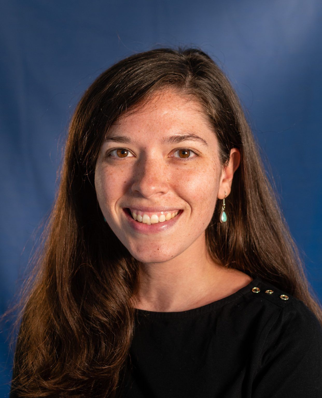A smiling woman with long brown hair wearing simple earrings in front of a blue background.