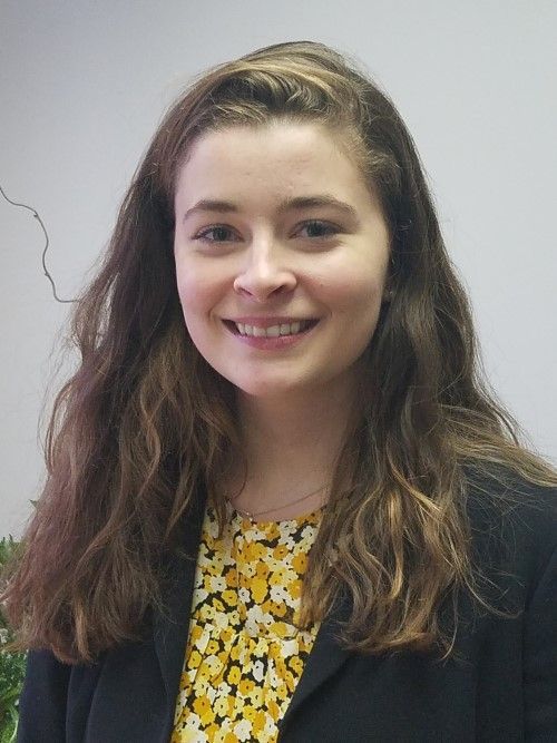 A smiling person with wavy brown hair, wearing a yellow floral top under a black blazer against a plain white wall.