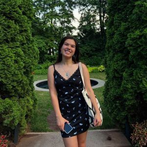 A smiling person in a black patterned slip dress and a tote bag stands between two tall hedges in an outdoor garden.