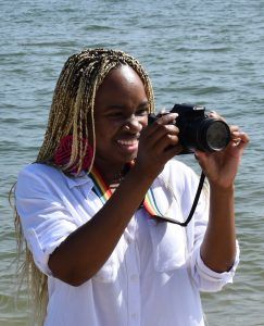 A person with braided hair wears a white shirt and rainbow lanyard while holding a camera to take a photo by the water.