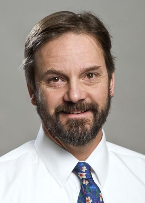 A professional headshot of a person with a beard, wearing a white collared shirt and a patterned blue tie.