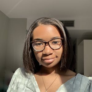 A person with dark hair and glasses poses indoors in natural, bright sunlight, wearing a light patterned top.