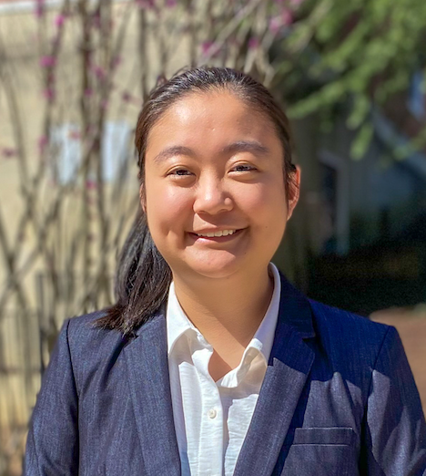A smiling person wearing a white collared shirt and dark blazer, standing outdoors with blurred branches in the background.