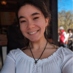 A smiling person wearing a white, off-the-shoulder top and a black pendant necklace, posed for a selfie in an outdoor area.