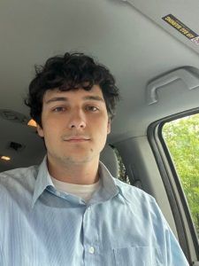 A person with curly dark hair wearing a light blue collared shirt, shown in a car interior.
