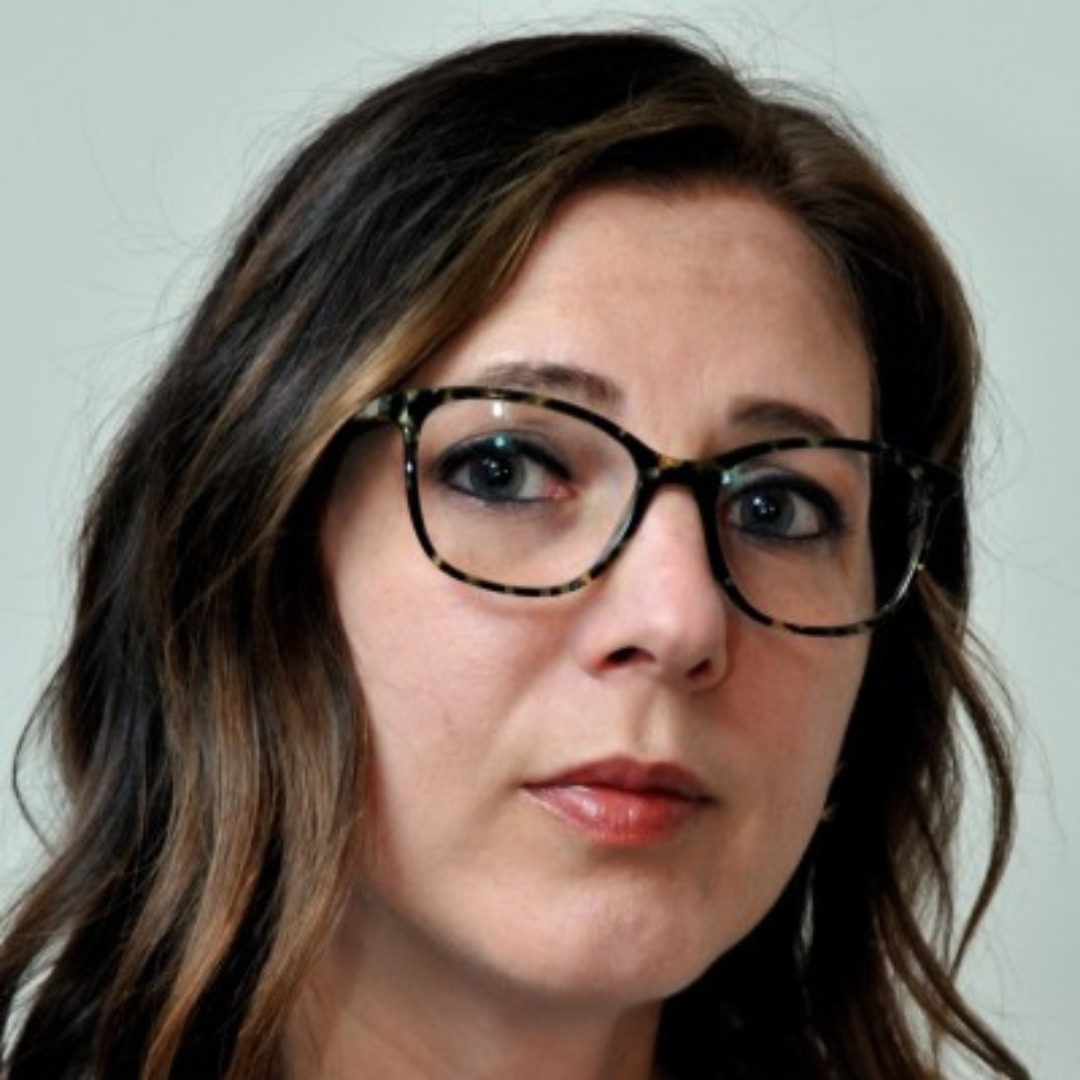 A close-up portrait of a person with wavy, dark hair wearing dark-rimmed glasses against a neutral background.