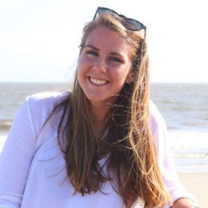 A person with long blonde hair and sunglasses on their head smiles at the camera on a beach with the ocean in the background.