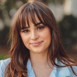 A person with long brown hair and bangs, smiling softly, wearing a light blue shirt in an outdoor setting.
