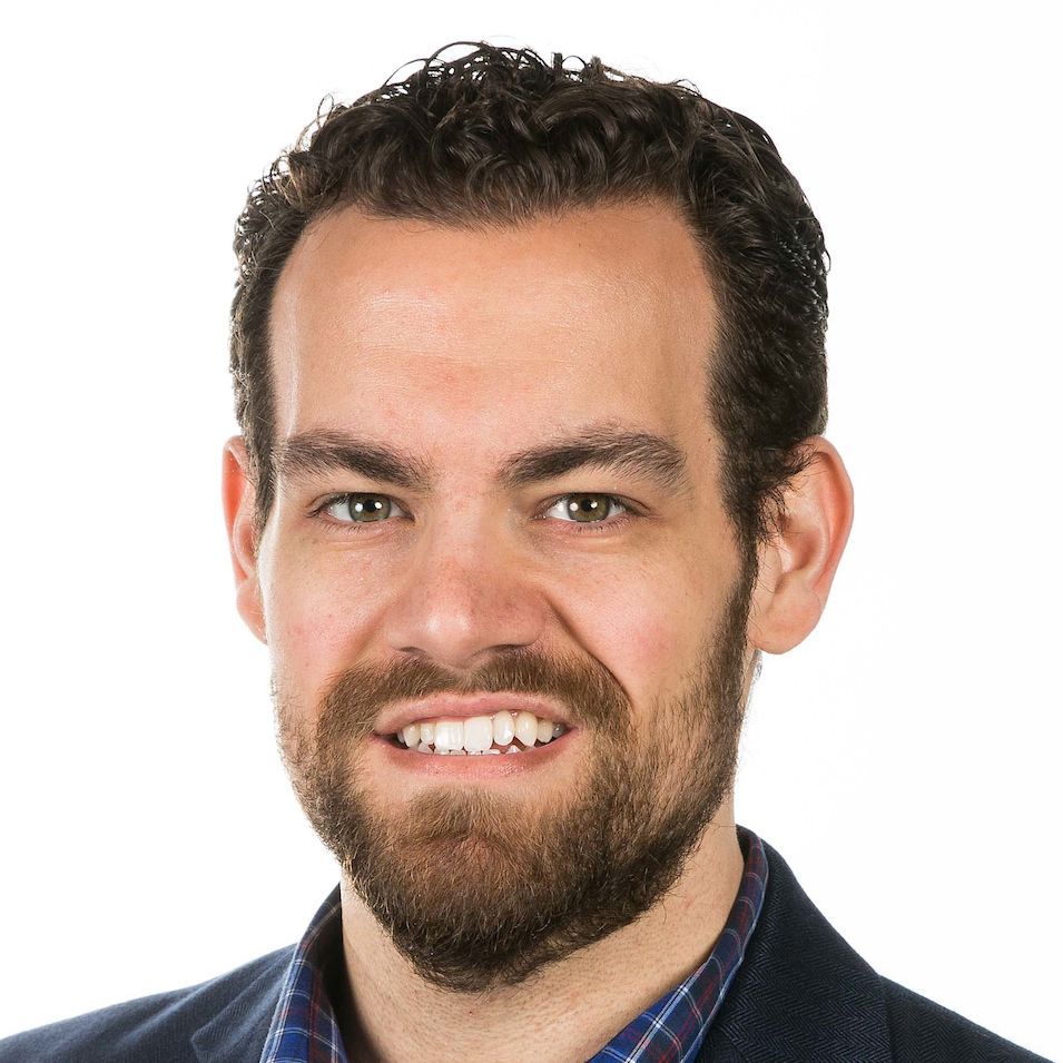 A professional headshot of a person with curly brown hair and a beard, wearing a navy blazer over a plaid shirt.