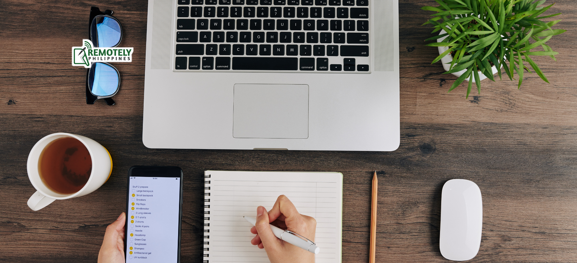 Desk with a laptop, smartphone, notepad with a hand writing, eyeglasses, a cup of tea, a plant, and a computer mouse.