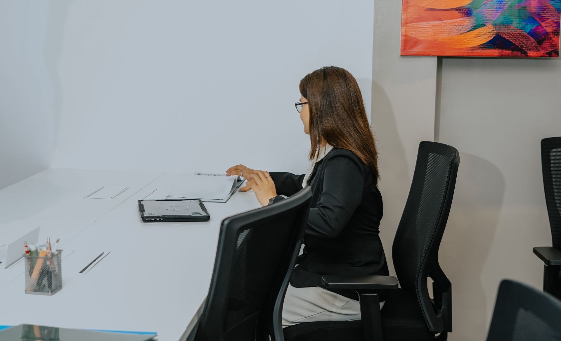 Woman in black blazer at a white desk, working with papers. Next to her is a black chair and tablet.