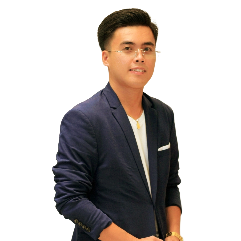 A person wearing a brown three-piece suit, white shirt, and striped tie with round glasses, smiling against a white backdrop.