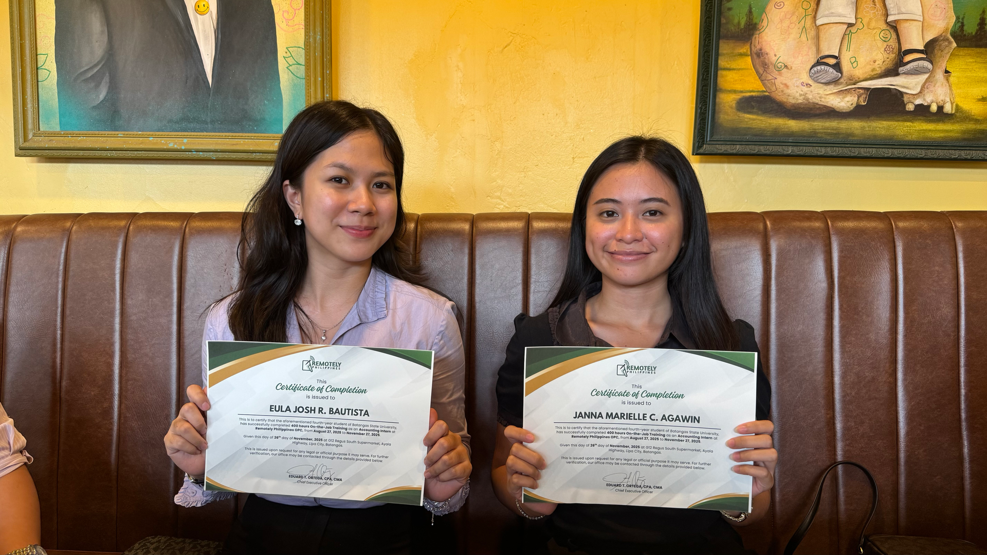 Two individuals sitting on a brown leather booth, smiling while holding up matching certificates in front of them.