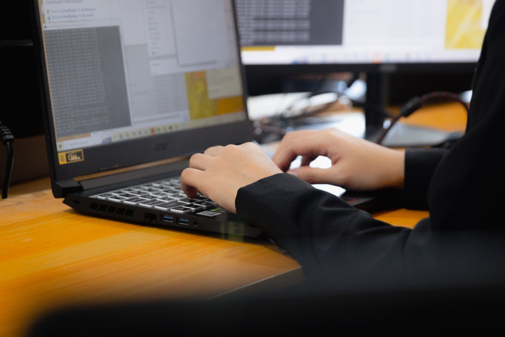 Person typing on a laptop with a second monitor in the background, set on a wooden desk.