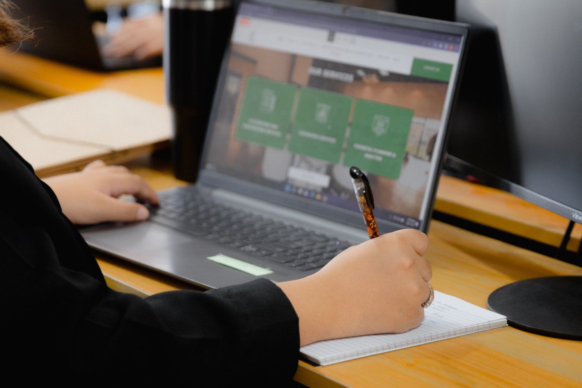 A woman is sitting at a desk using a laptop computer and writing in a notebook.