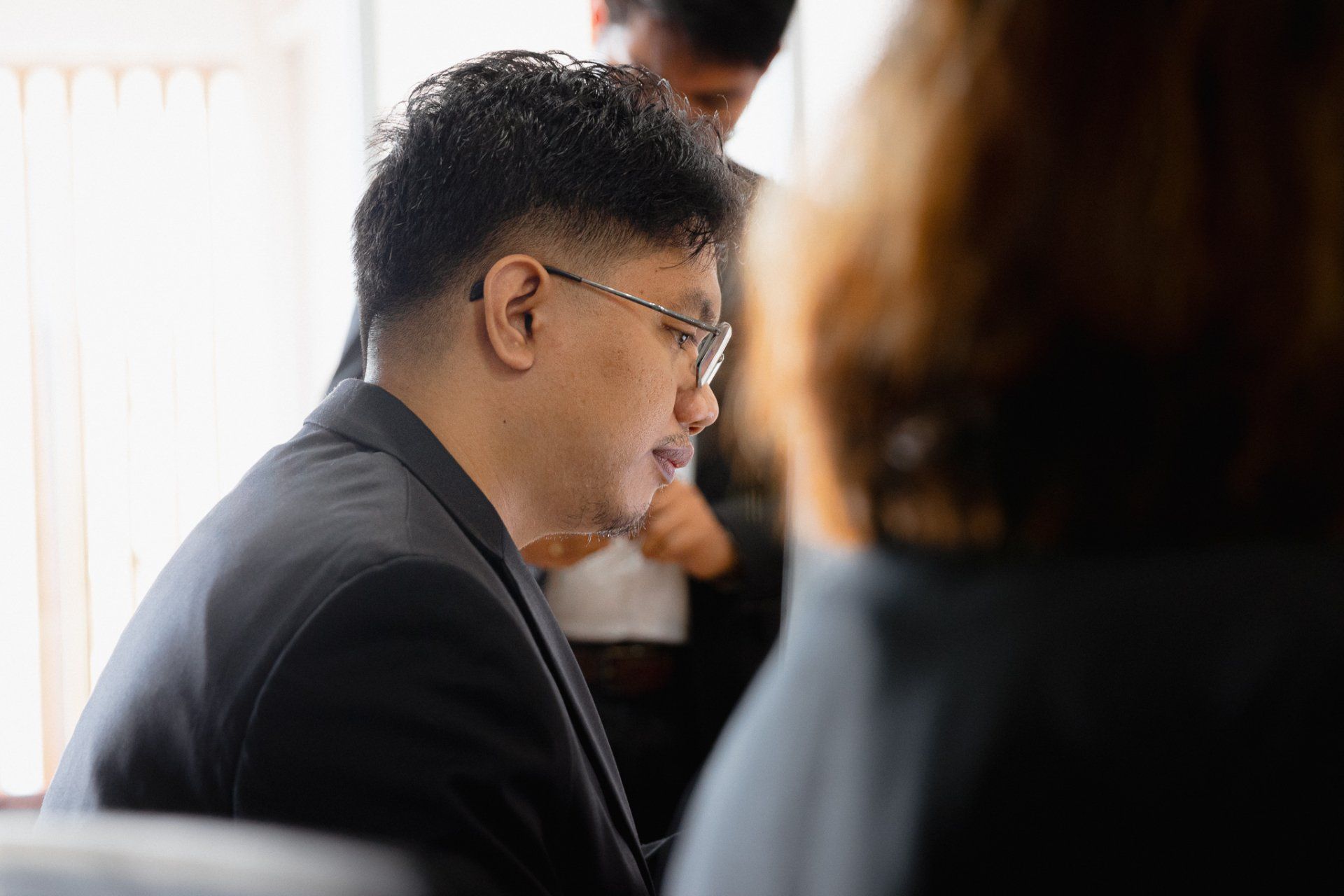 Man in a black suit with glasses; indoors, seated, head turned to the side, looking down.