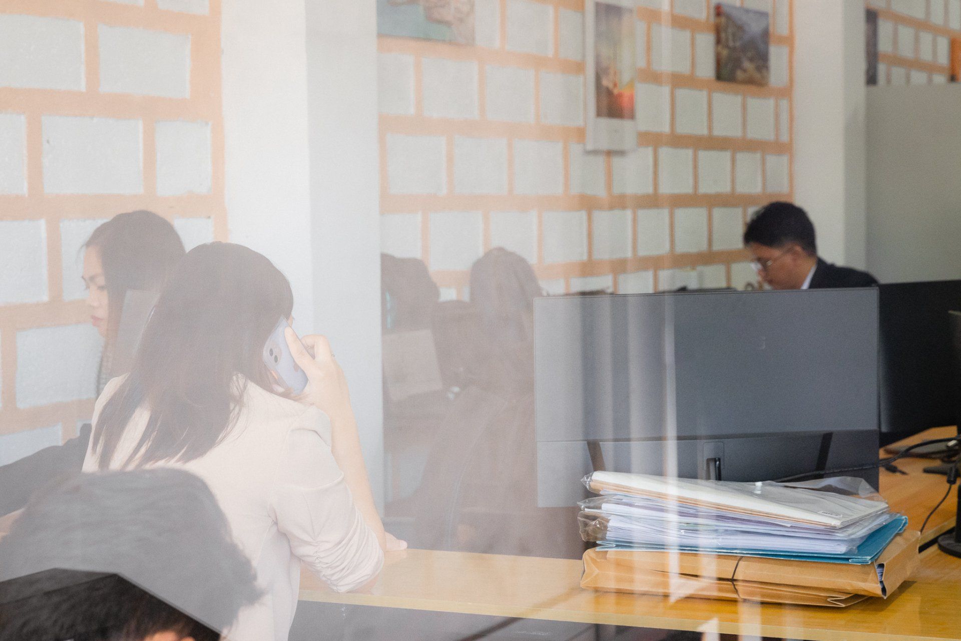 Office setting. People near computer monitors and desks. Documents and pictures on walls.