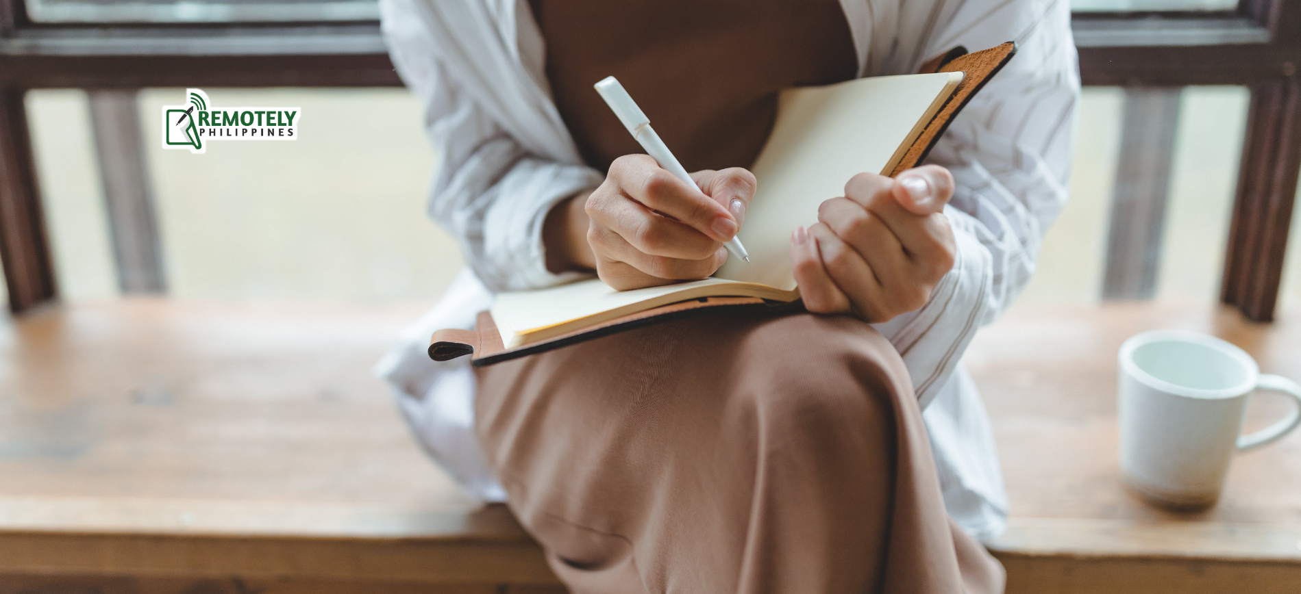 Person writing in a notebook beside a white mug on a wooden table.