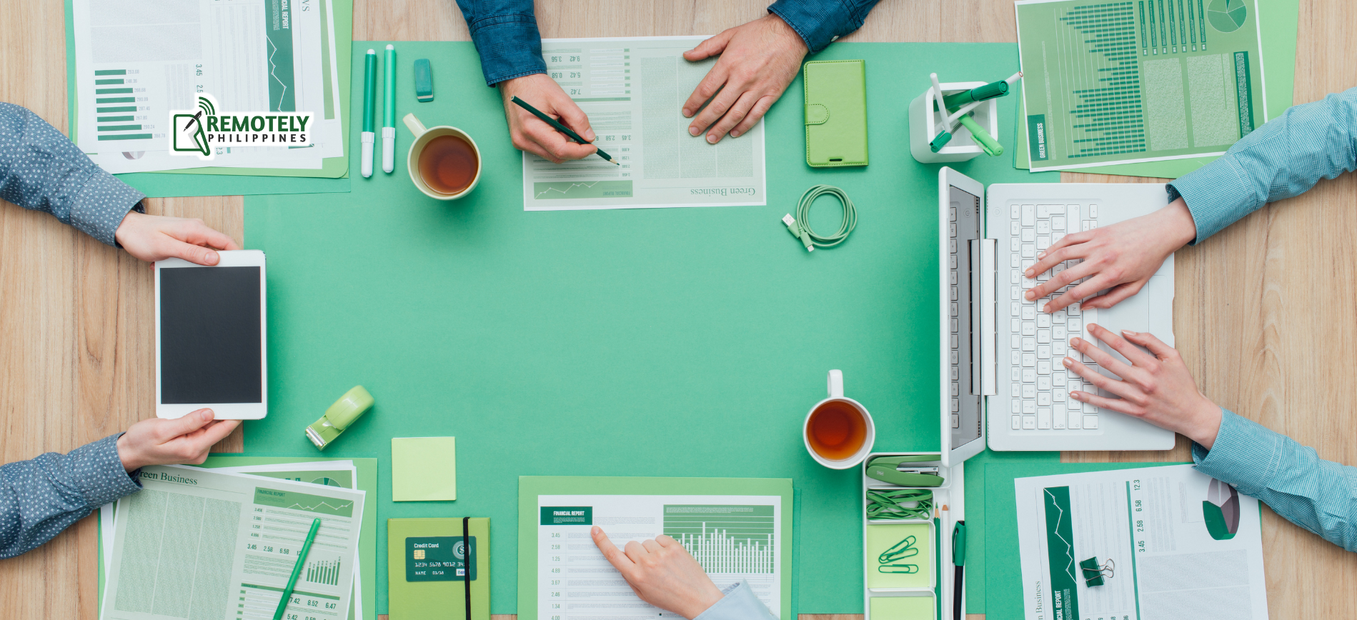 Overhead view of a meeting table with documents, coffee cups, hands, a tablet, and a laptop