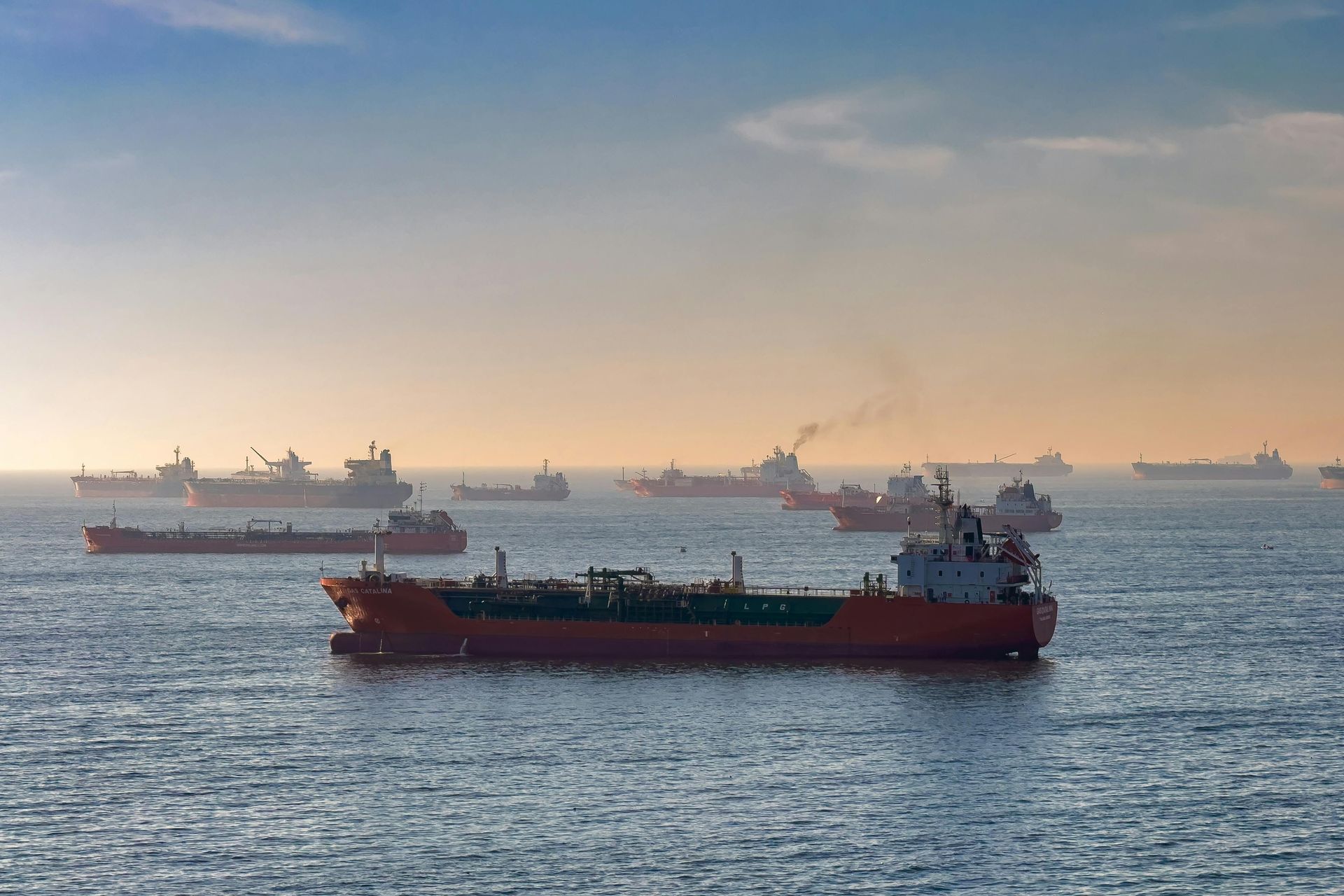 Cargo ship on calm sea at sunrise, with several distant ships on the horizon and a hazy sky.