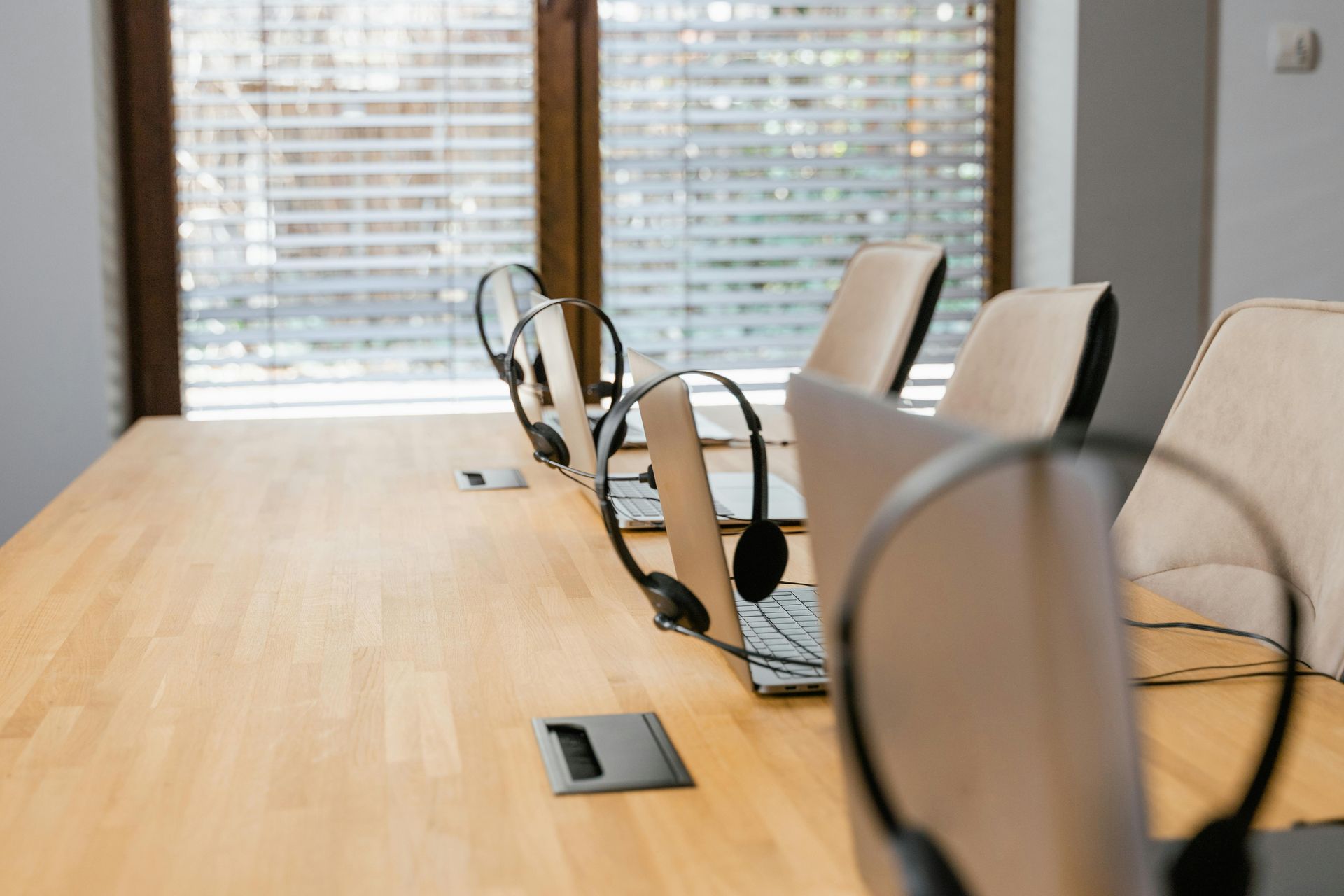 Conference room with a long wooden table, chairs, and laptop docks by a window with blinds