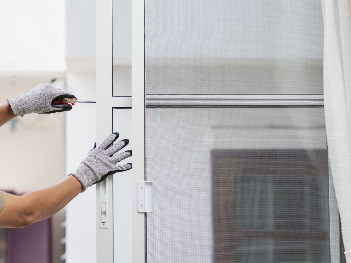 Gloved hands adjusting a white window frame beside a screened window