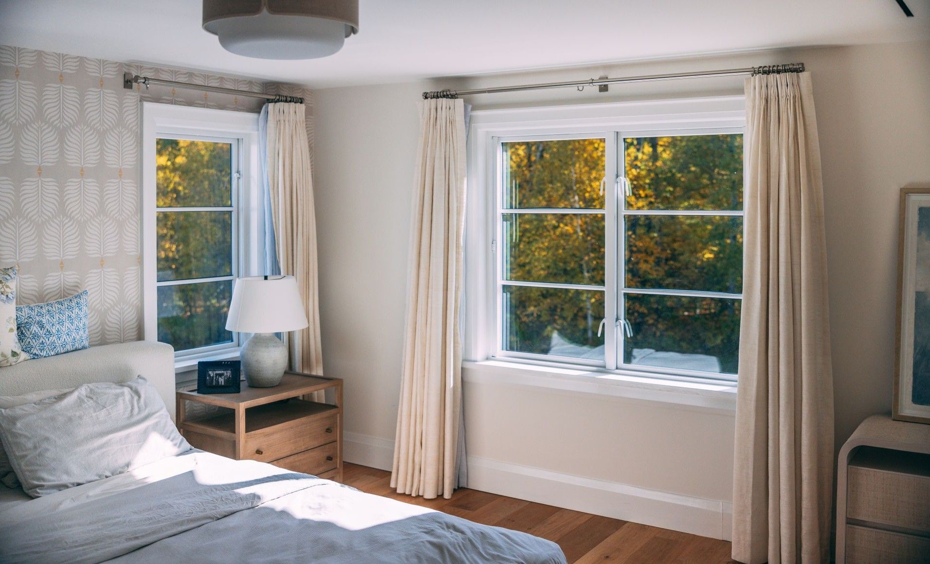 Bright bedroom with bed, nightstand, lamp, and a large window showing autumn trees