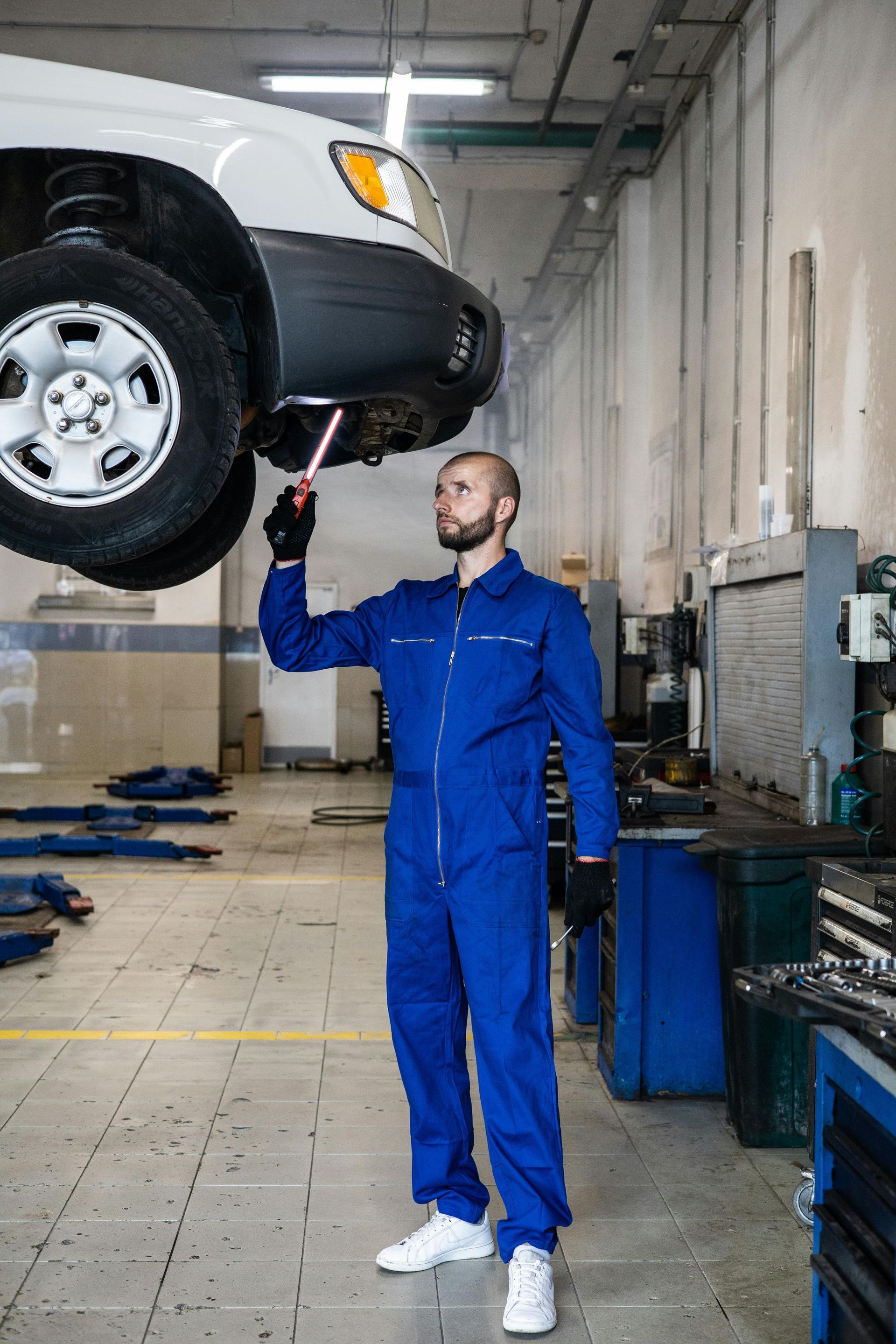 Mechanic in blue jumpsuit examining car lifted on a hoist in a garage.