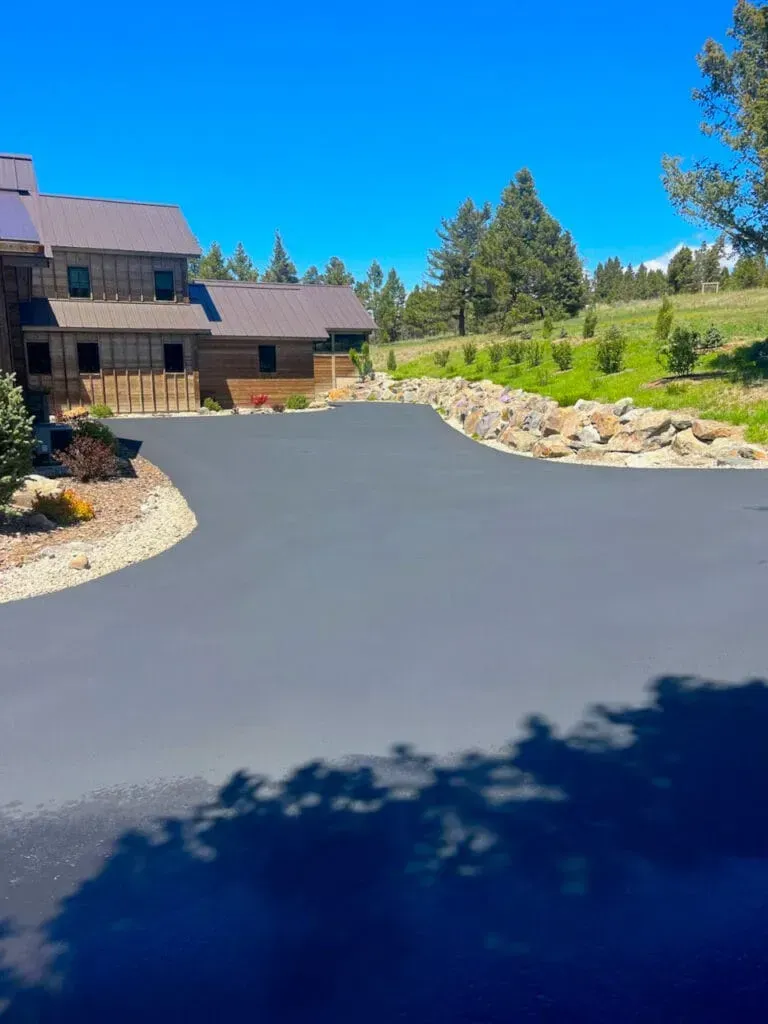 Dark asphalt driveway leading to a wood-sided house under a blue sky, with trees and a grassy hillside.