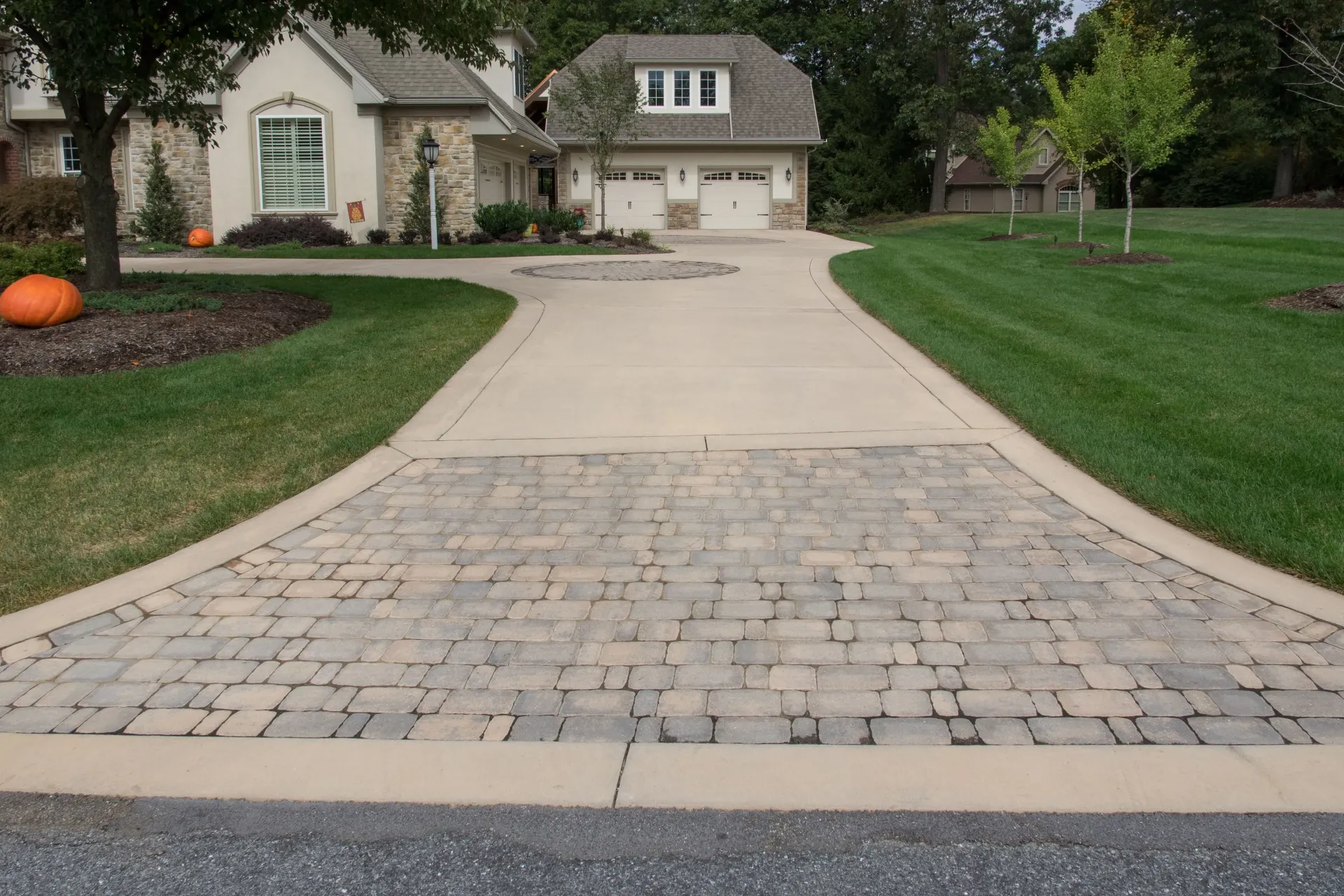 Paver driveway leading to a house with a garage, green lawn, and trees.