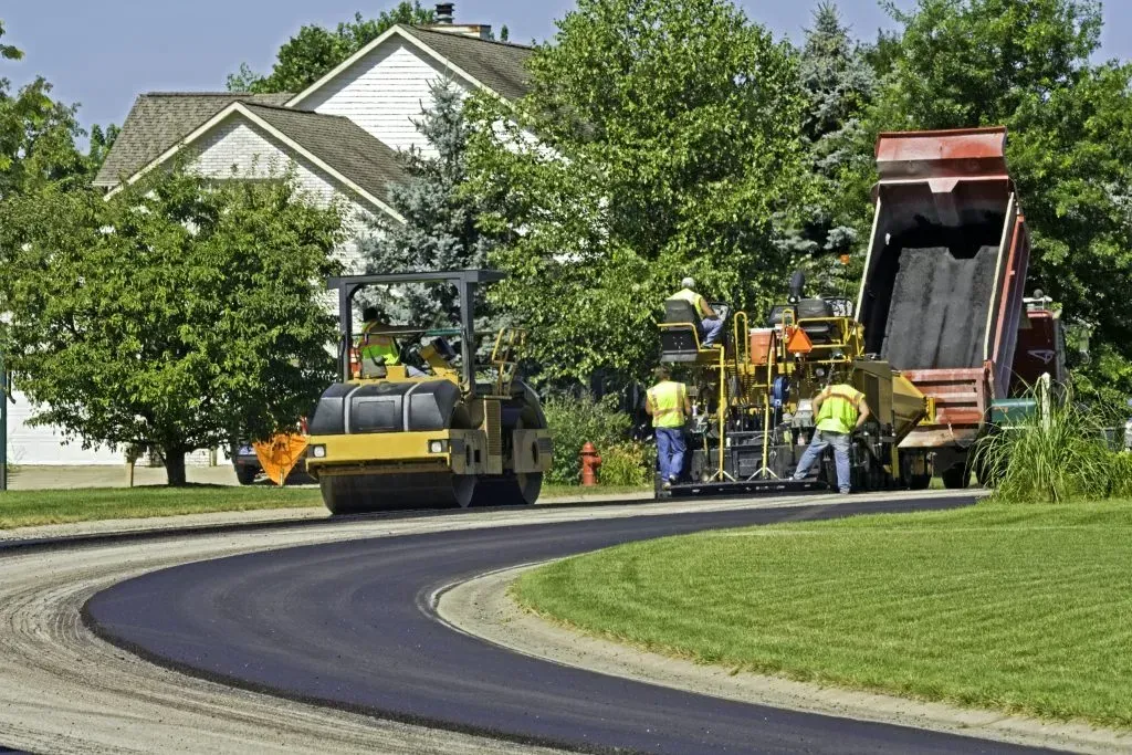 Road paving in progress, featuring asphalt, workers, machinery, and a residential backdrop.