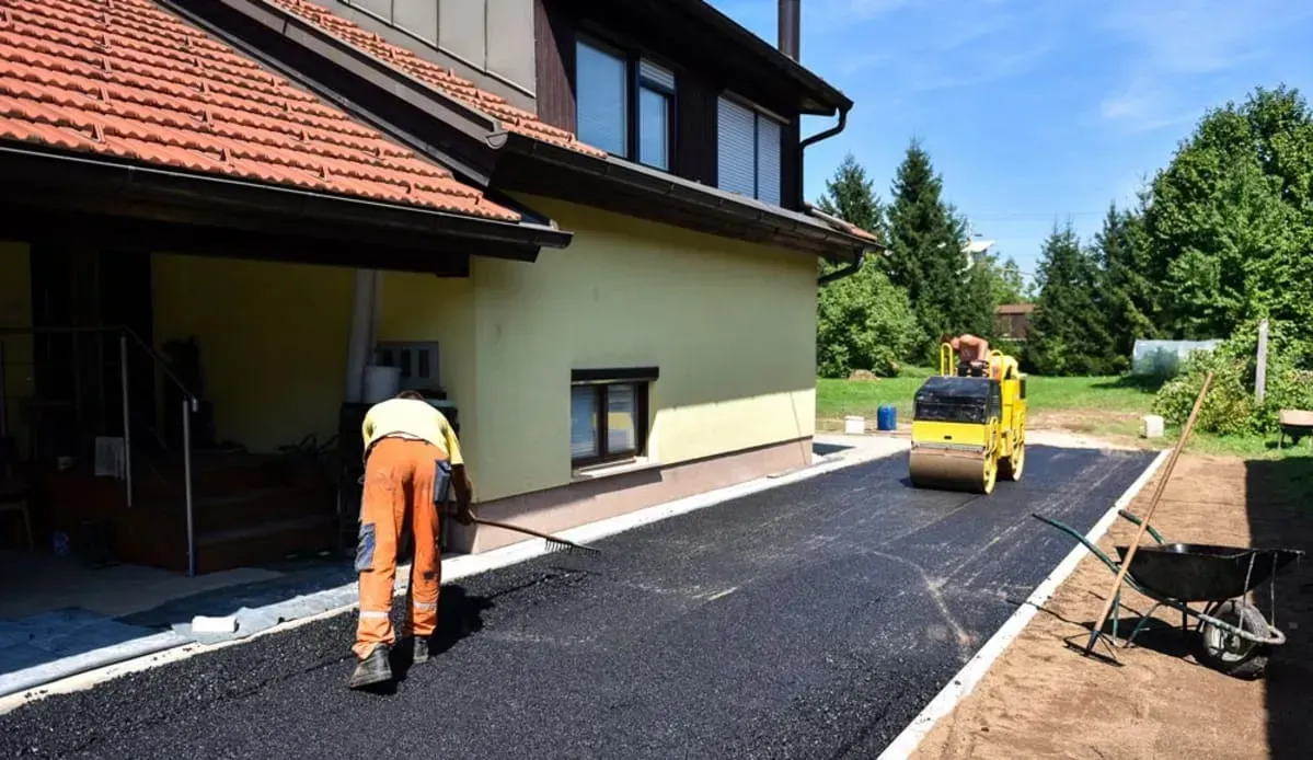Asphalt paving of a driveway, near a house. Worker in orange pants and a small roller flatten the surface.
