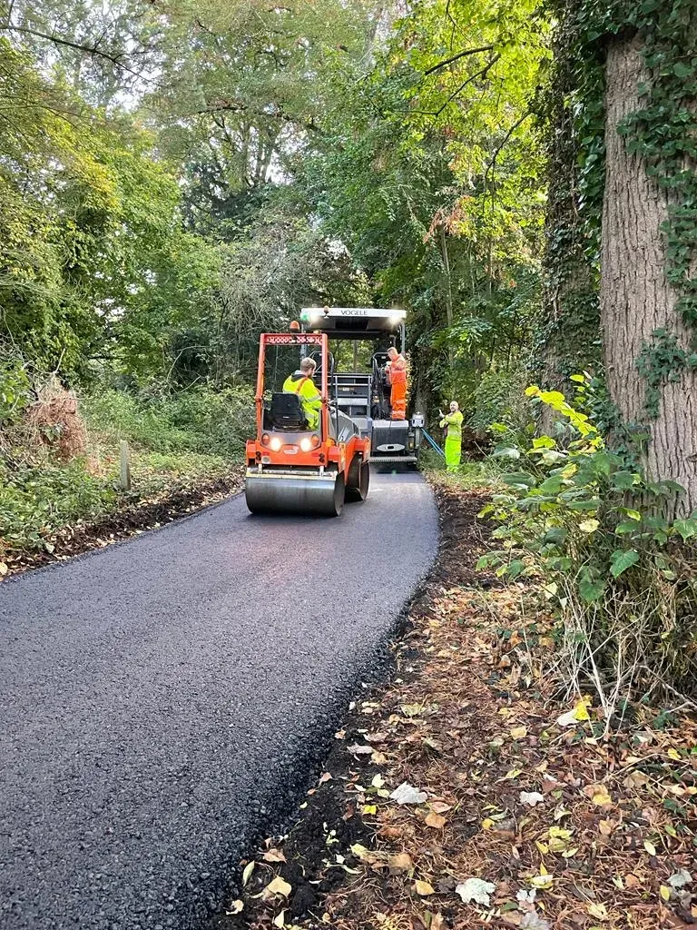 Road paving in progress; orange roller compacting asphalt. Worker in yellow vest nearby, surrounded by trees.