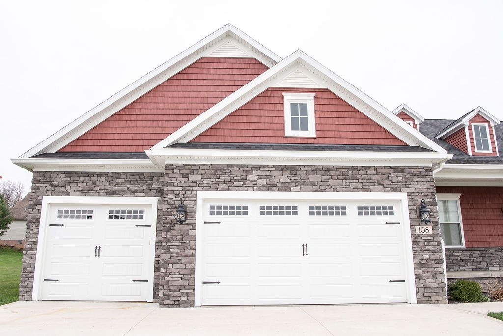 A red house with two white garage doors and a stone wall.