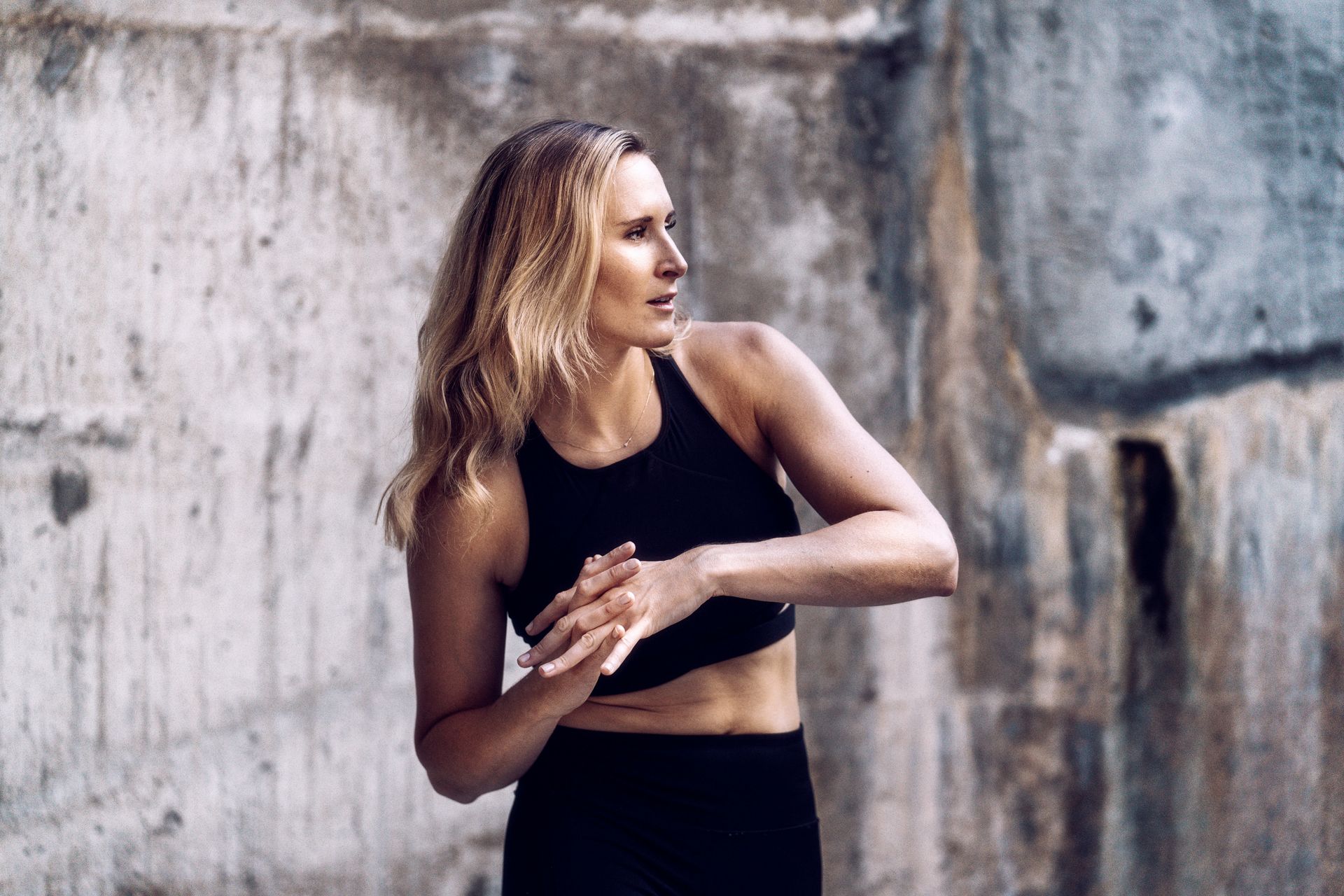 Woman in a black athletic outfit stretching beside a weathered concrete wall