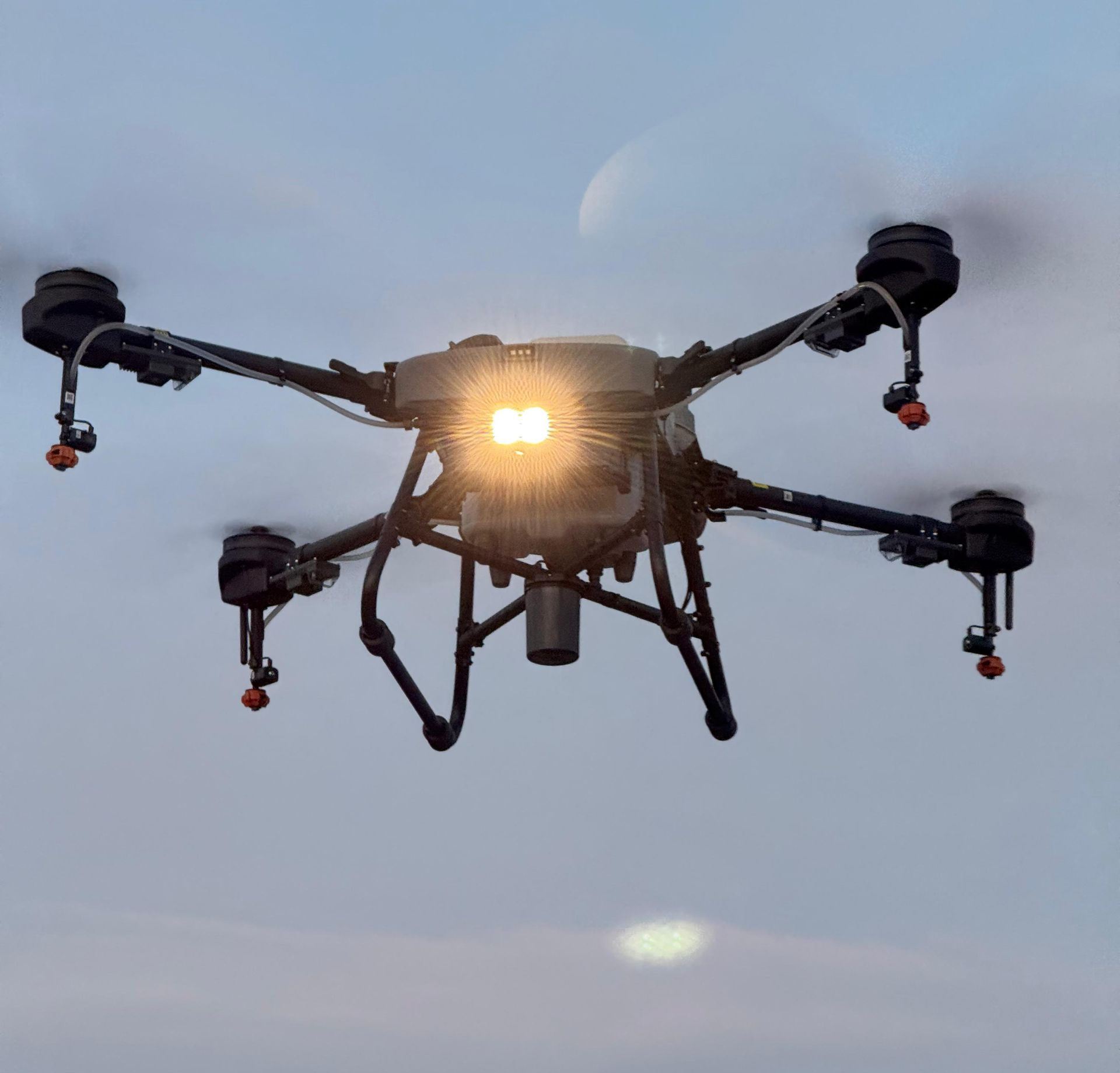 Dron volando sobre un campo verde con un bosque y montañas al fondo bajo un cielo azul.
