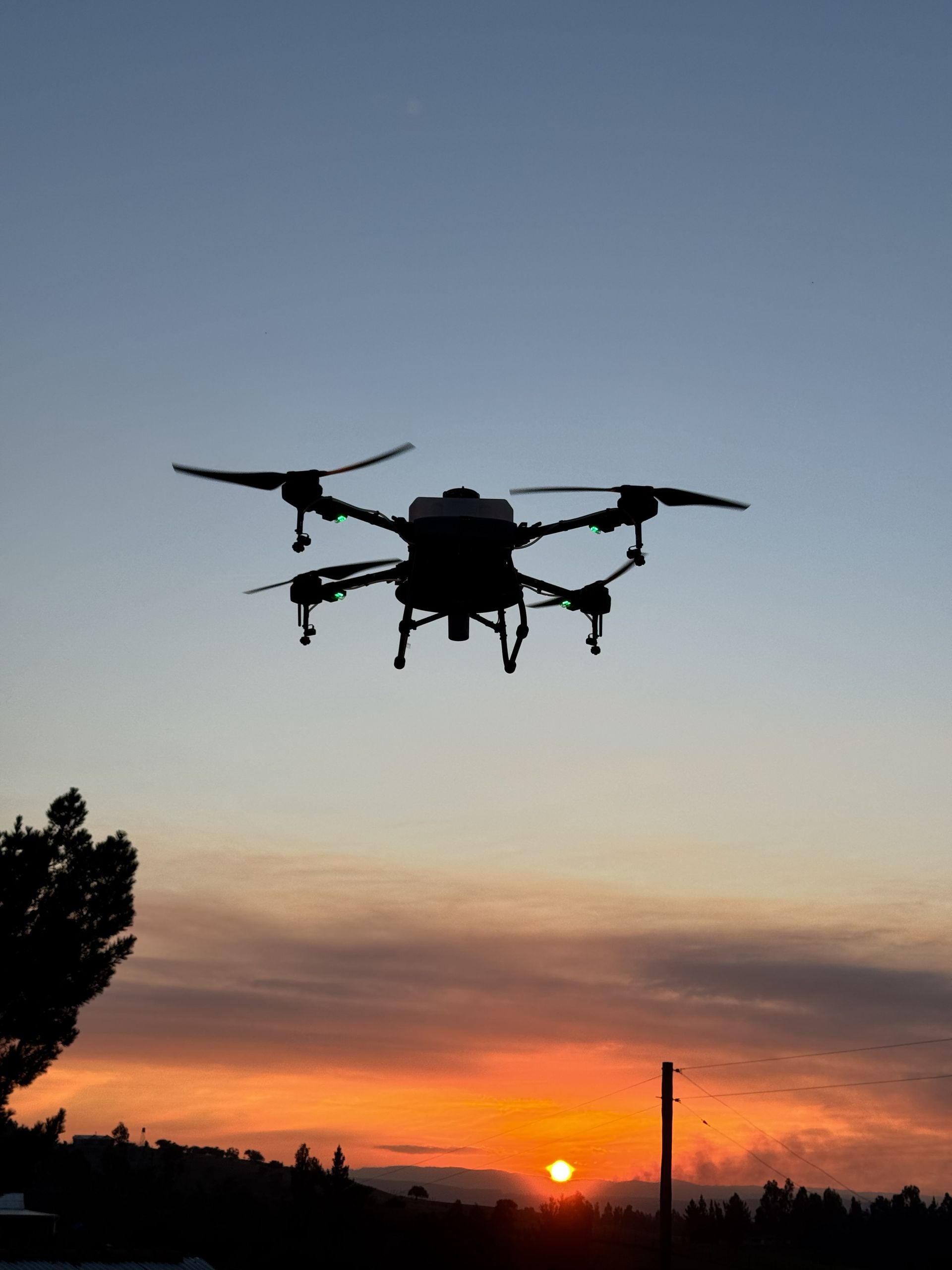 Dos drones agrícolas sobre tierra roja frente a un campo y montañas bajo un cielo nublado.