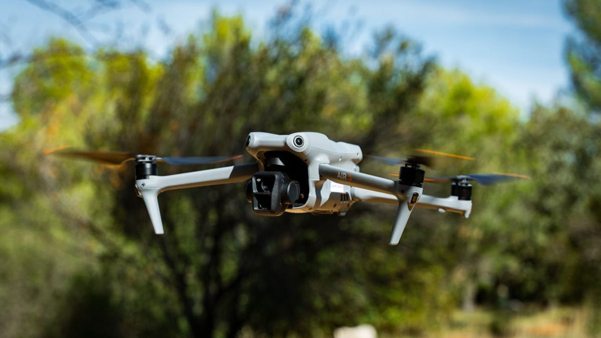 Dron blanco con cámara negra en vuelo; árboles verdes borrosos en el fondo.