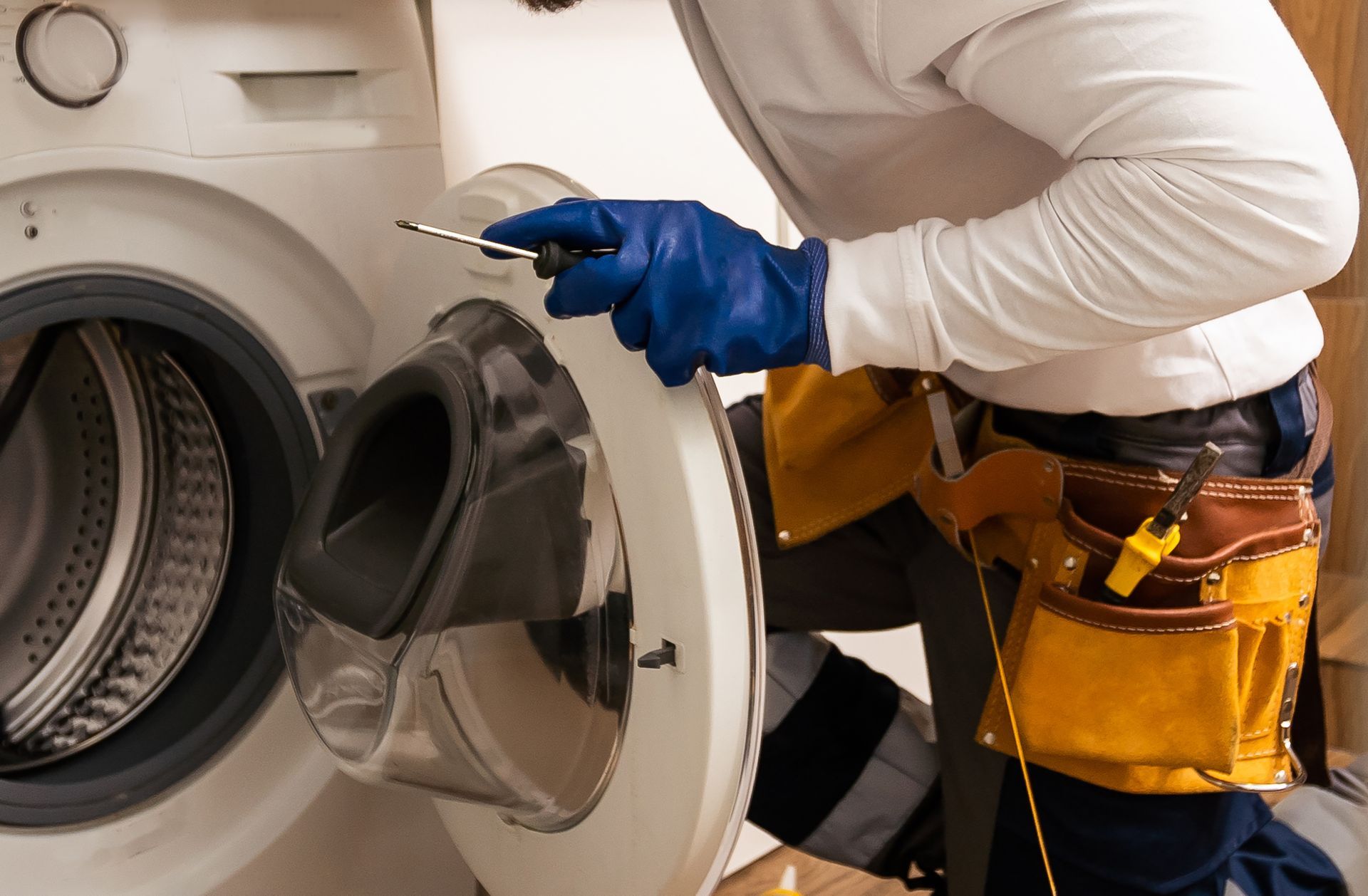 Technician in blue gloves inspecting an open front-loading washing machine with a tool belt.