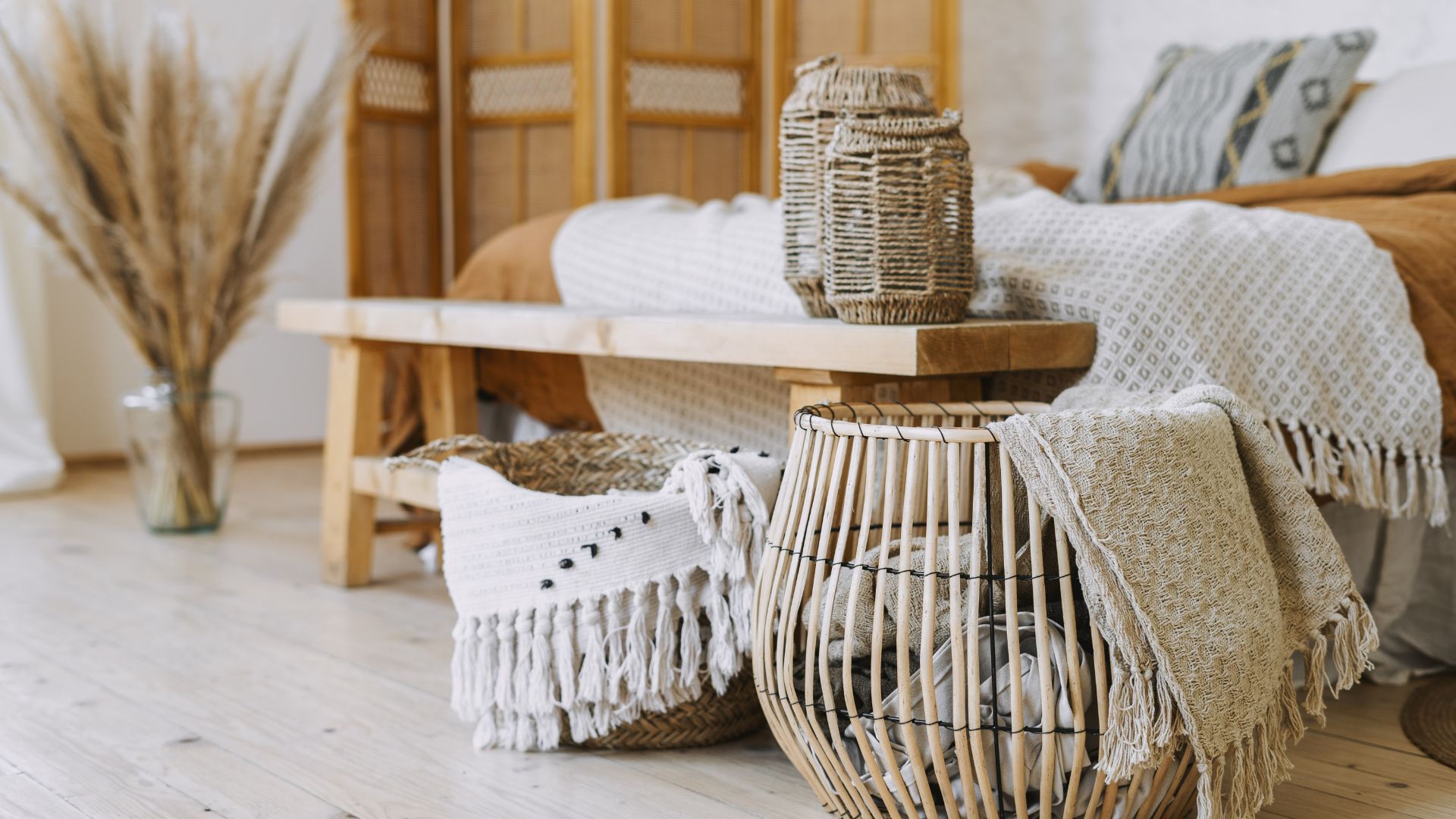 A bedroom with a bed , a wooden bench , a wicker basket and a vase of dried flowers.