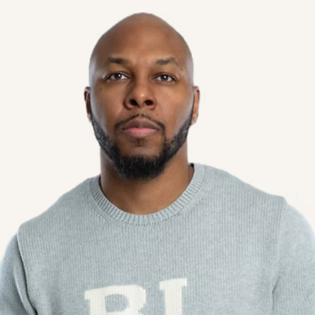 A bald man with a beard is wearing a grey sweater with the letter r on it