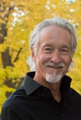 A man with gray hair and a beard is wearing a black shirt and smiling.