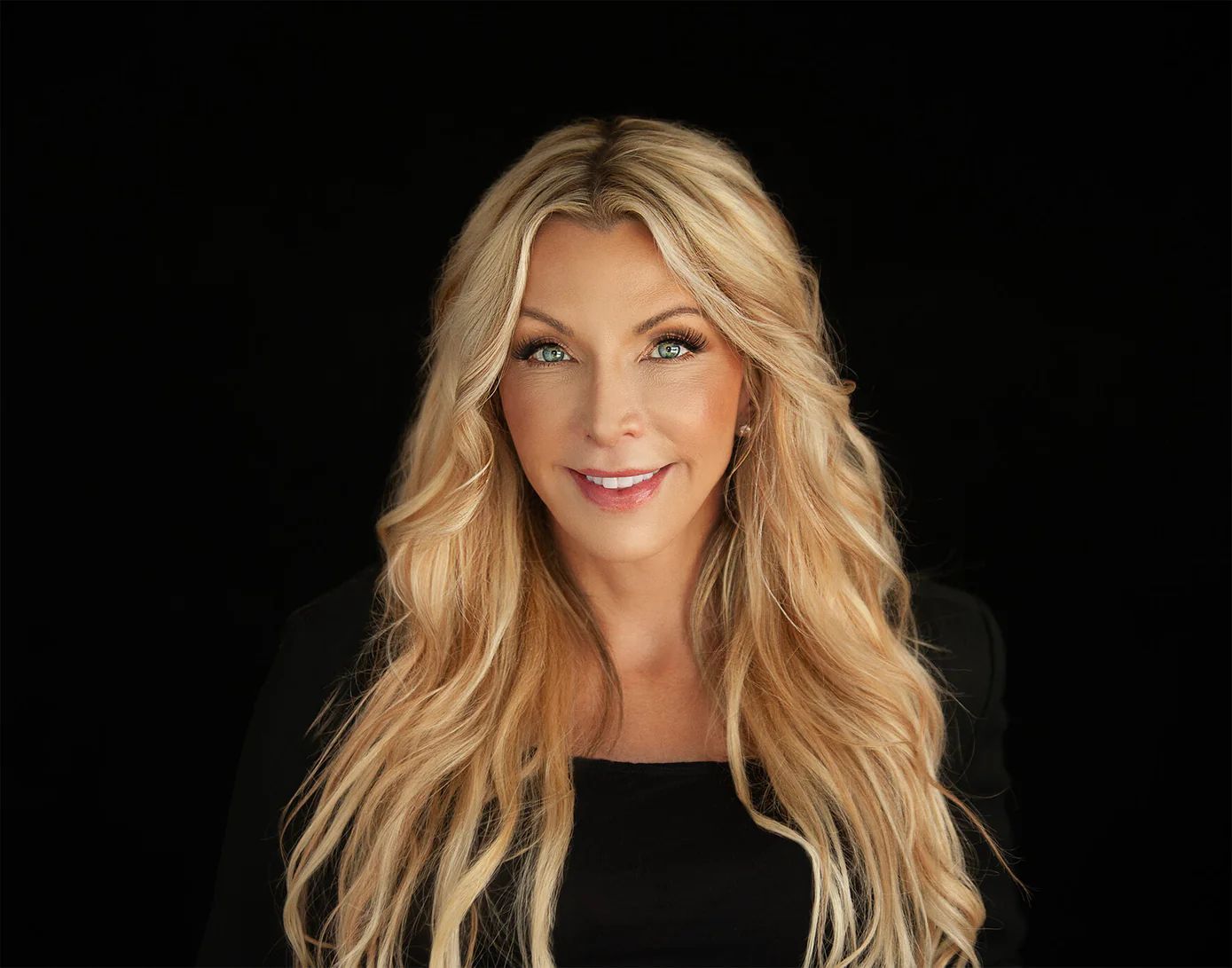 A woman with long blonde hair is wearing a black dress and smiling.