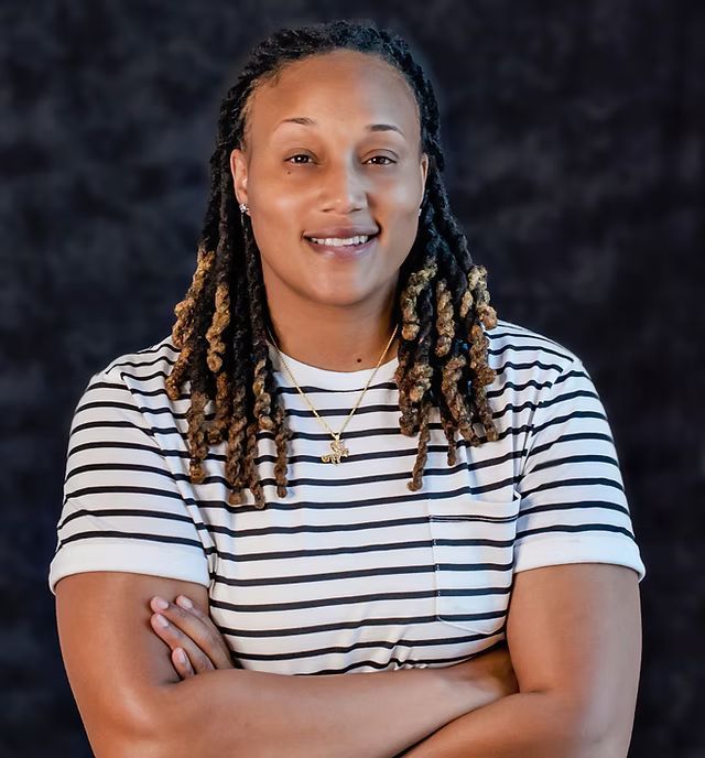 A woman with dreadlocks is wearing a striped shirt and smiling with her arms crossed.