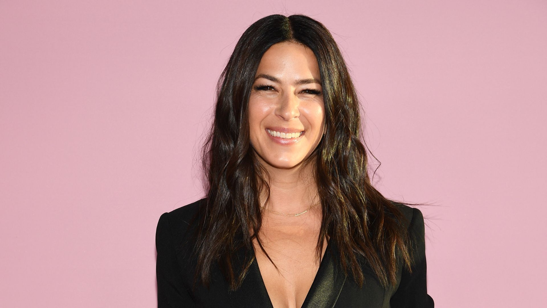 A woman wearing a black jacket and a plunging neckline is smiling on a pink background.
