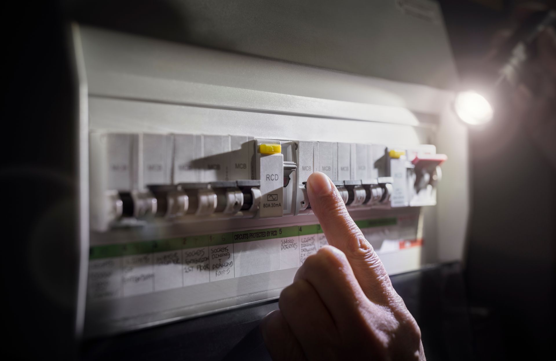 Man in a dark room using a flashlight to turn on or off a circuit breaker on an electrical fuse board during a power outage emergency.