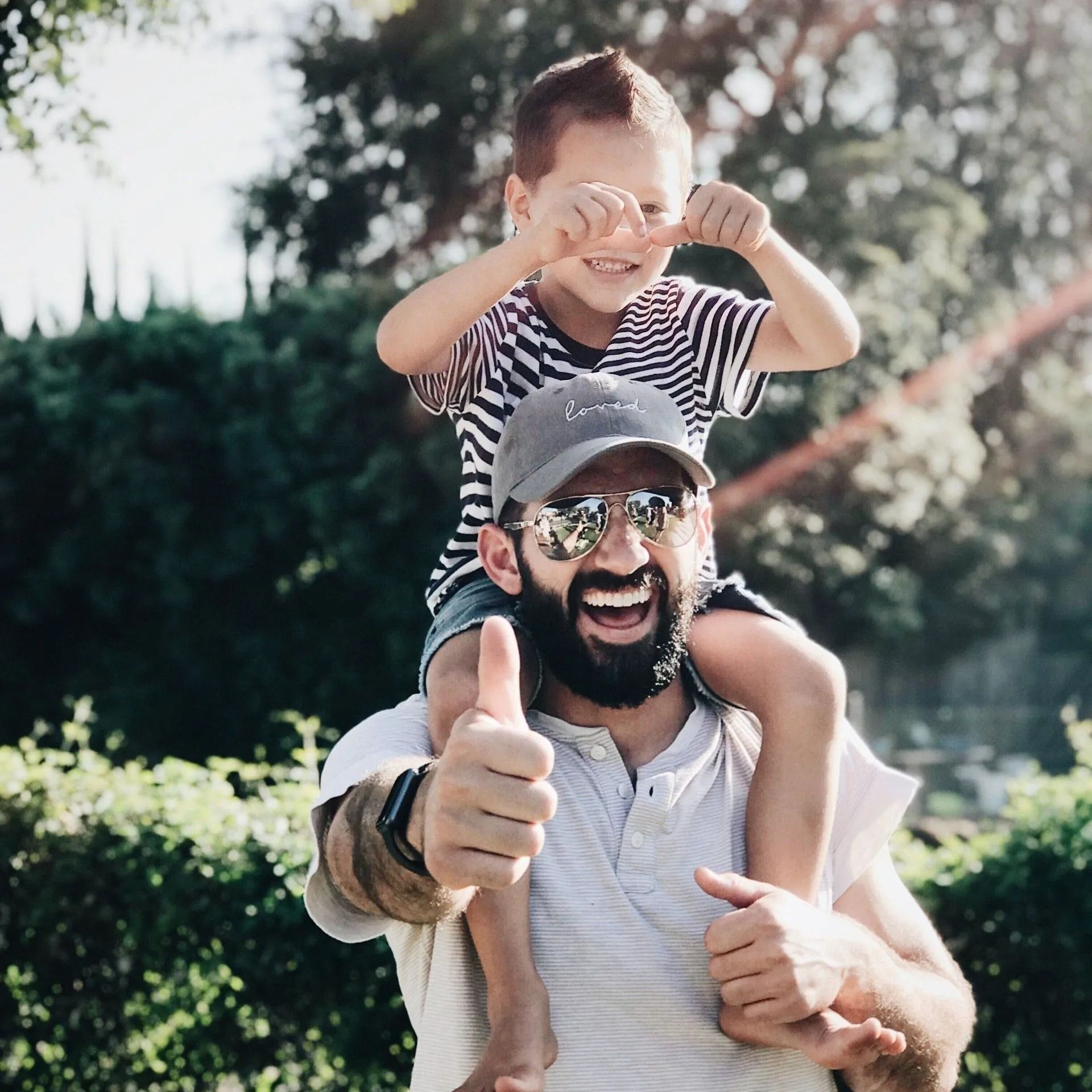Smiling man giving a piggyback ride to a child outdoors, both wearing striped shirts and hats.