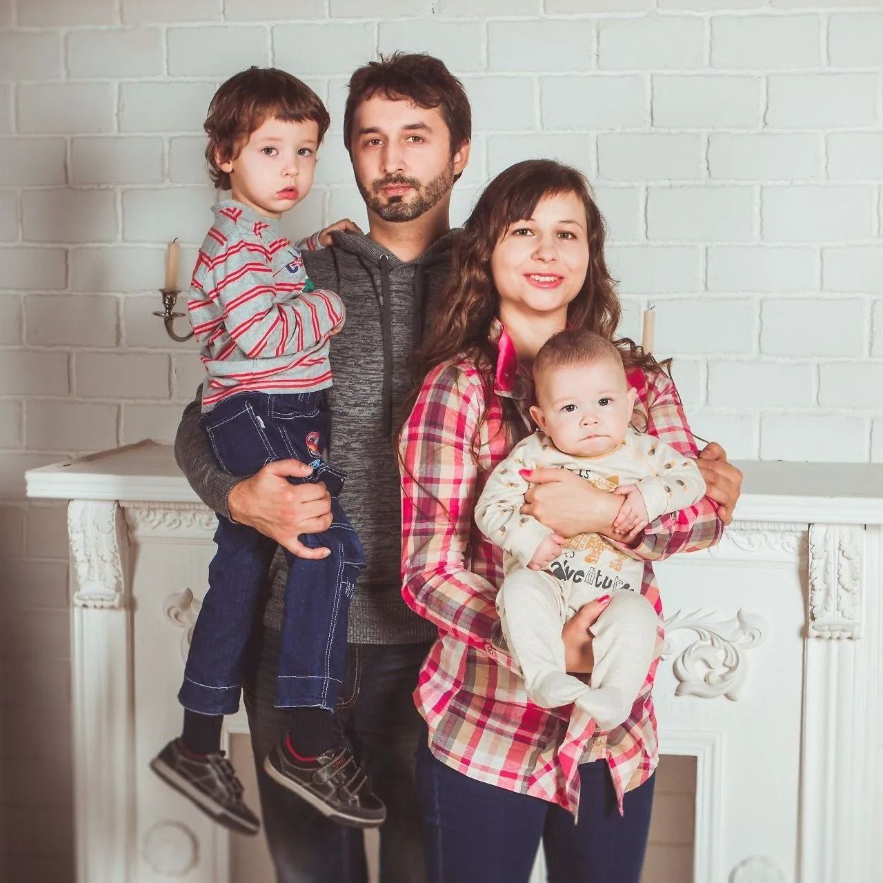 Family portrait of four posing in front of a white brick wall and mantel, holding two young children.