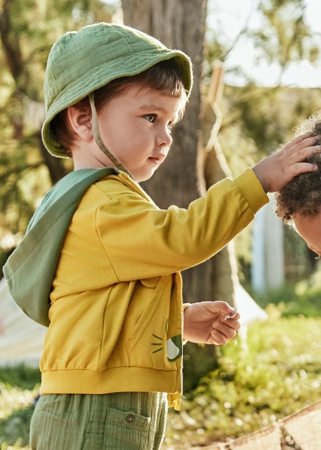 Un ragazzo con la giacca gialla e il cappello verde tocca la testa di un altro all'aperto, in una giornata di sole.