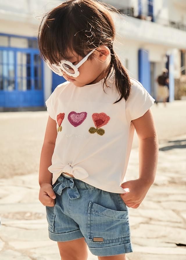 Ragazza con occhiali da sole, maglietta annodata sul davanti con disegni floreali, pantaloncini di jeans, in piedi all'aperto con le mani in tasca.